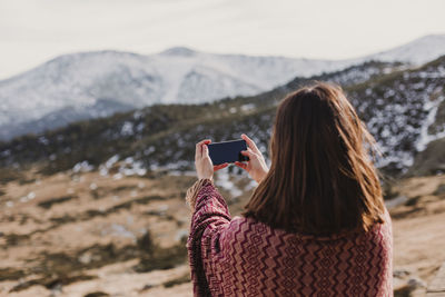 Rear view of woman photographing on mobile phone against mountains and sky