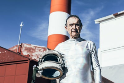 Low angle of senior male astronaut with helmet looking at camera while standing against shabby buildings of spaceport on sunny day