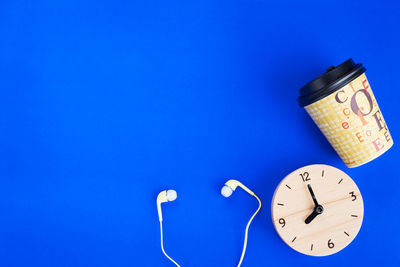 High angle view of clock with disposable cup and headphones over blue background