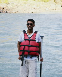 Young man wearing life jacket while standing against river