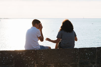 Rear view of friends sitting on retaining wall against sea during sunset