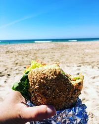 Cropped image of hand holding  a sandwich roll at beach against sky 
