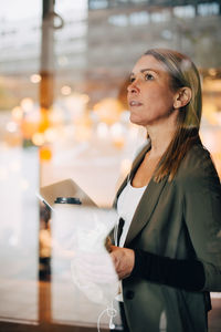 Mature businesswoman holding food and drink with laptop standing in cafe seen from window