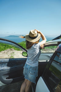 Young woman driver holds straw hat on head and admires the seascape. person is hitchhiking in wild