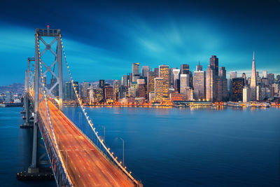 Illuminated city by river against blue sky