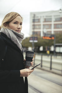 Businesswoman holding smart phone and disposable coffee cup at tram station