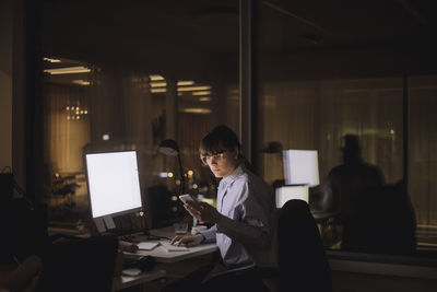 Businesswoman text messaging on mobile phone sitting in office at night