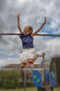 Low angle view of girl with arms raised jumping against cloudy sky