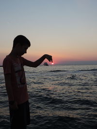 Optical illusion of silhouette boy touching sun against sea during sunset