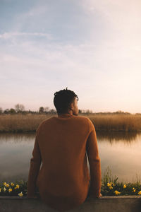 Rear view of man sitting by lake against sky during sunset