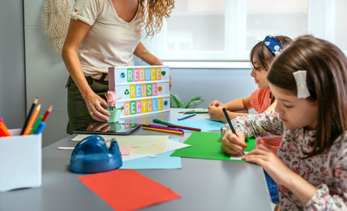 Teacher holding lightbox with text reduce, reuse, recycle while students drawing in classroom