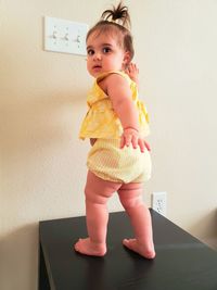 Cute baby girl looking away standing on table against wall at home