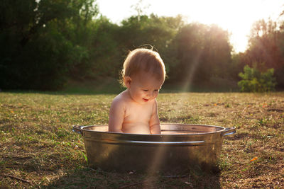 Cute little kid sits in a basin of water in nature and has rays of the setting sun, golden bathe. 