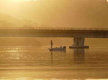 Man fishing in river by bridge during sunset