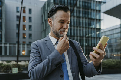 Thoughtful mature businessman looking at smart phone in front of building