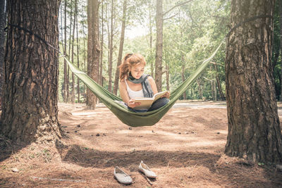 Front view of woman reading book while sitting on hammock in forest