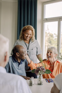 Smiling young female caregiver standing by happy senior women and men at dining table in nursing home