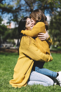 Side view of mother embracing daughter while kneeling on grassy field in park