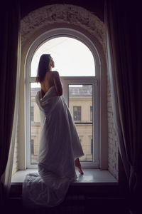 Woman in a white blanket is standing on a white windowsill with her back to him