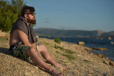 Bearded man sitting at beach against sky