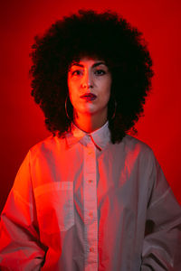 Unemotional young ethnic female model with dark afro hair in white shirt standing against red background and looking at camera