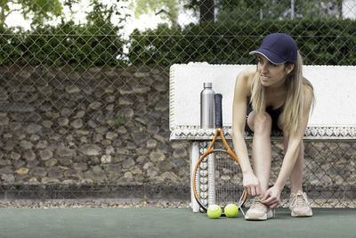 Portrait of female tennis player tying shoelaces sitting on a bench in a tennis court copy space
