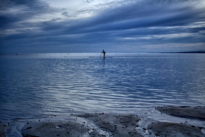 Distant view of person paddleboarding in sea against cloudy sky