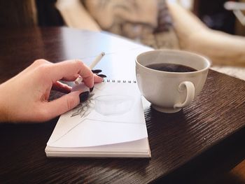 Cropped hand of woman with drawing in book while having coffee at table