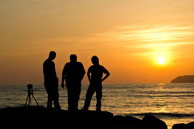 Silhouette friends standing on cliff by sea against sky during sunset