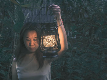 Mature woman holding illuminated string lights in lantern at forest