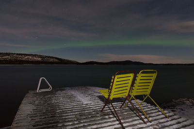 Chairs on snow covered pier at river against sky with aurora borealis