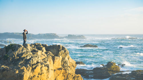 Man standing on rock by sea against sky