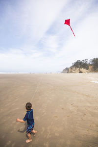 Young boy looks up at red kite flying in the sky at the beach.