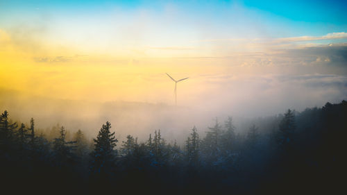 A wind turbine behind a forest covered in fog during a colorful sunset.