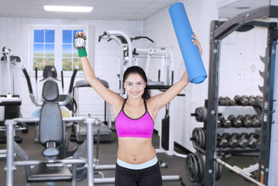 Portrait of confident mid adult woman holding clock and exercise mat while standing in gym