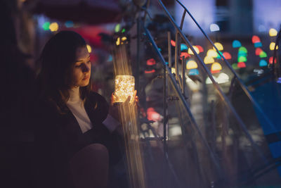 Thoughtful woman holding glowing jar while sitting on steps at night