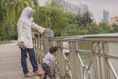 Side view of mother with son looking at lake while standing on footpath against trees
