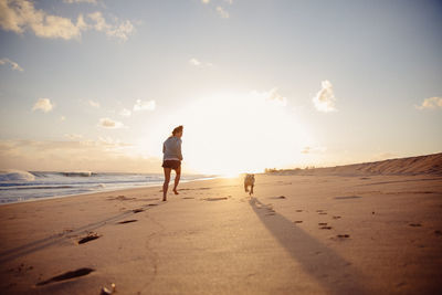 Rear view of woman with dog running at beach against sky