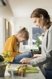 Woman cutting fruits with girl sitting on kitchen counter at home