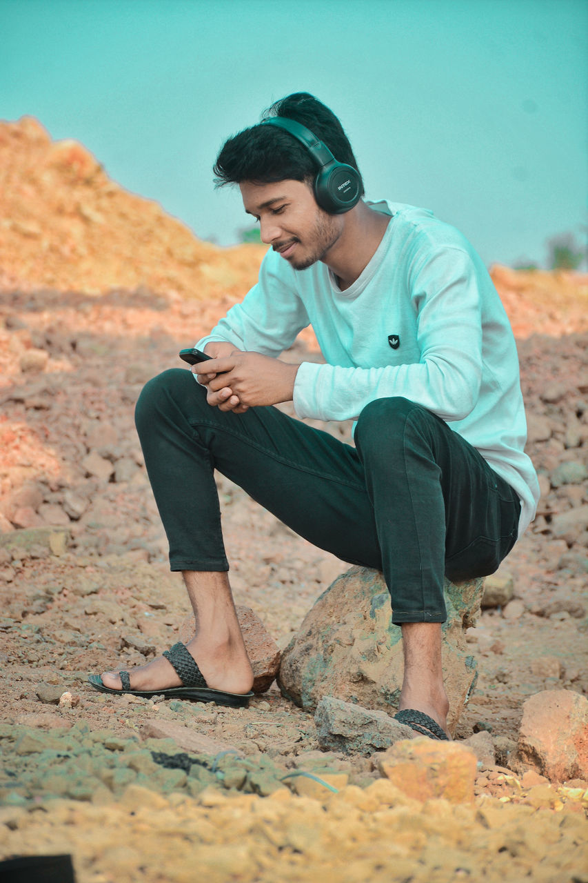 YOUNG MAN LOOKING AWAY WHILE SITTING ON ROCKS