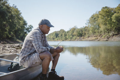 Man adjusting fishing tackle while sitting on boat by lake against clear sky