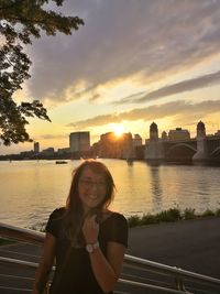 Portrait of smiling woman by river against sky during sunset
