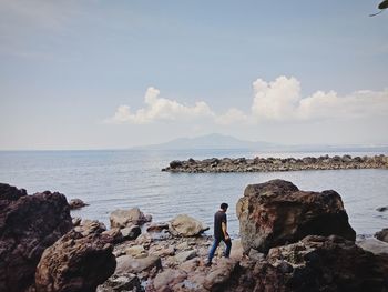 Side view of young man walking on rocks at beach against sky during sunny day
