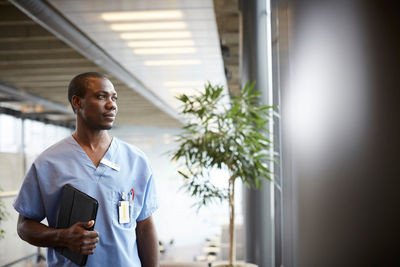 Thoughtful mid adult male nurse with digital tablet looking through window while standing in corridor at hospital