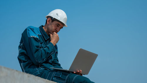 Man using laptop while standing against clear blue sky