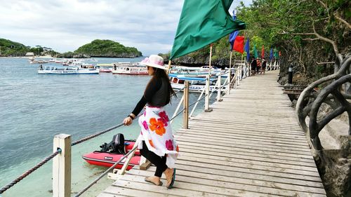 Woman on footbridge by sea with boats moored against sky