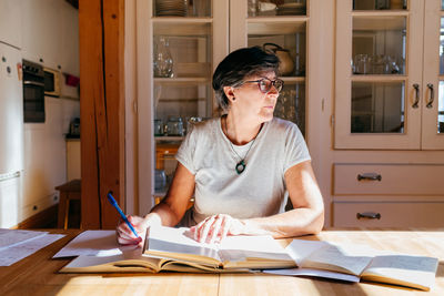 Focused middle aged female in glasses with pen in hand sitting at table with heap of books and papers and studying professional materials at home