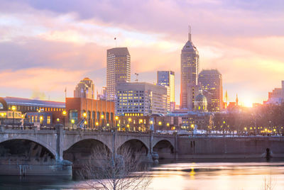 Bridge over river by buildings against sky during sunset