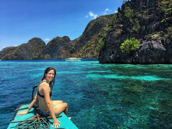 Young woman sitting by sea against blue sky