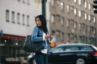 Teenage girl looking away while holding smart phone against building in city